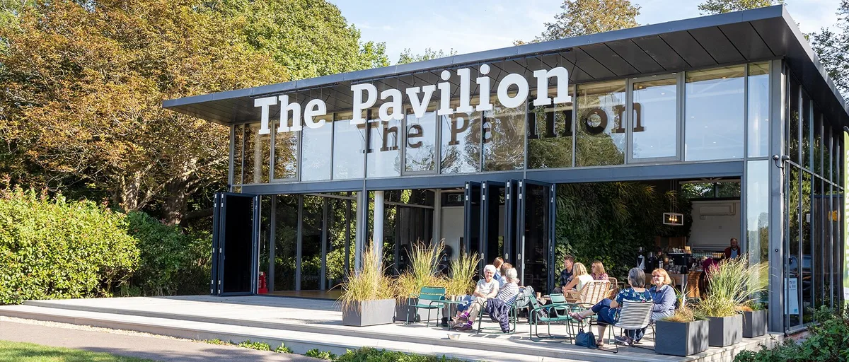 The Pavilion at Graylingwell Park, a glass-walled building with its name in large white letters, with people sitting at tables on the terrace outside.