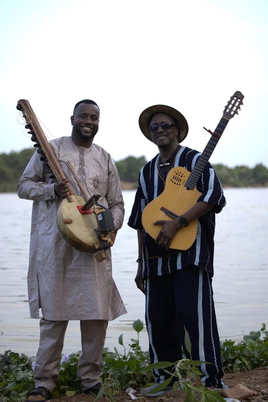 Sura Susso holding a kora and Habib Koité holding an acoustic guitar, standing side by side on a riverbank