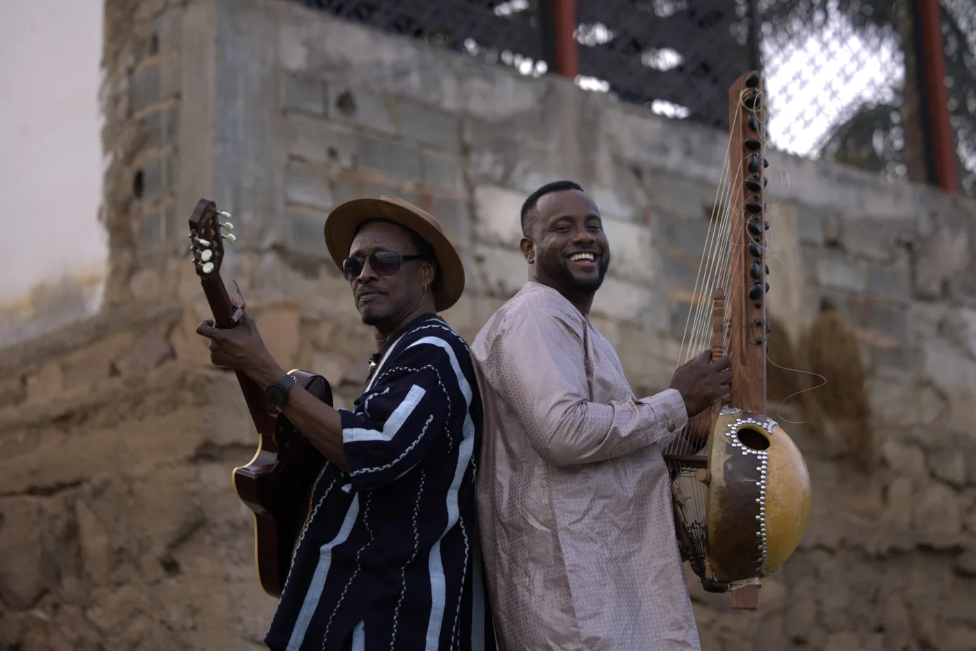 Habib Koité, holding a guitar, and Sura Susso, holding a kora, back to back against a stone wall