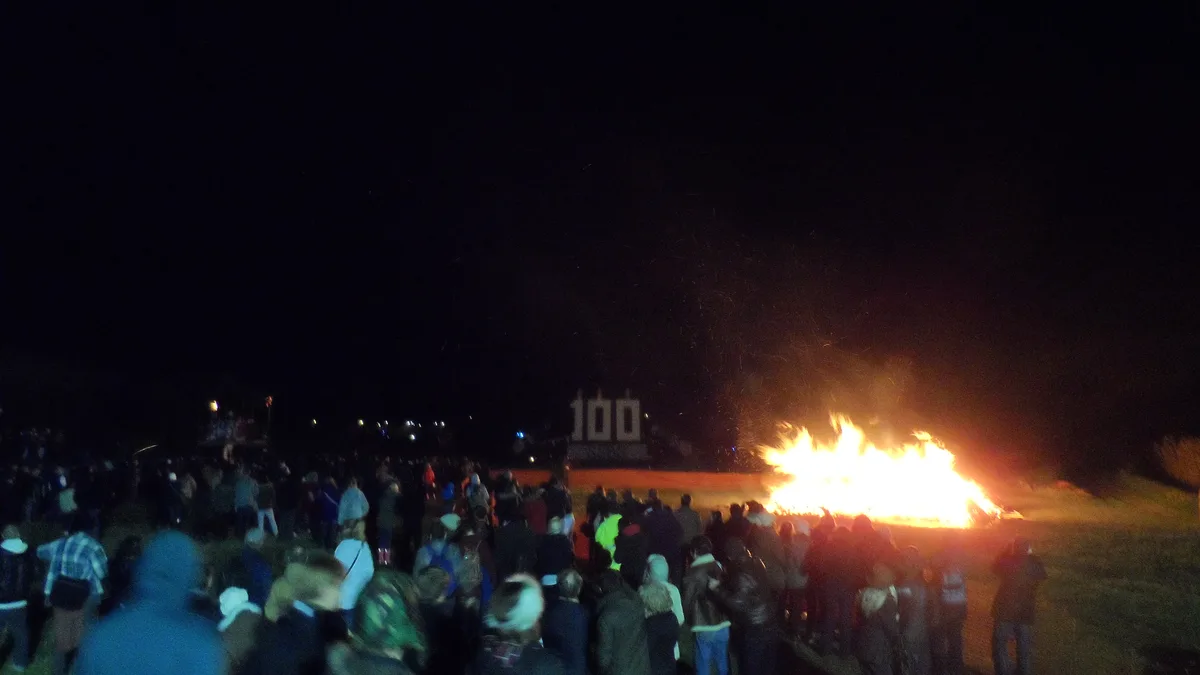 A crowd watching a large bonfire burn on open ground at night