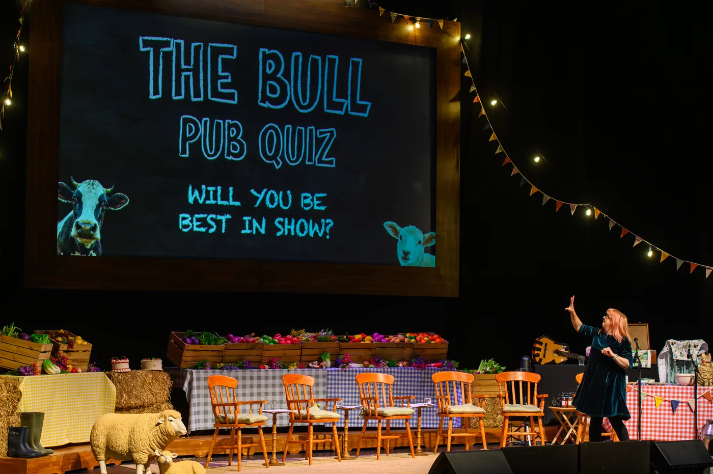 A large screen reading The Bull Pub Quiz, Will you be best in show?, above a stage of chairs and produce tables with a performer pointing at it