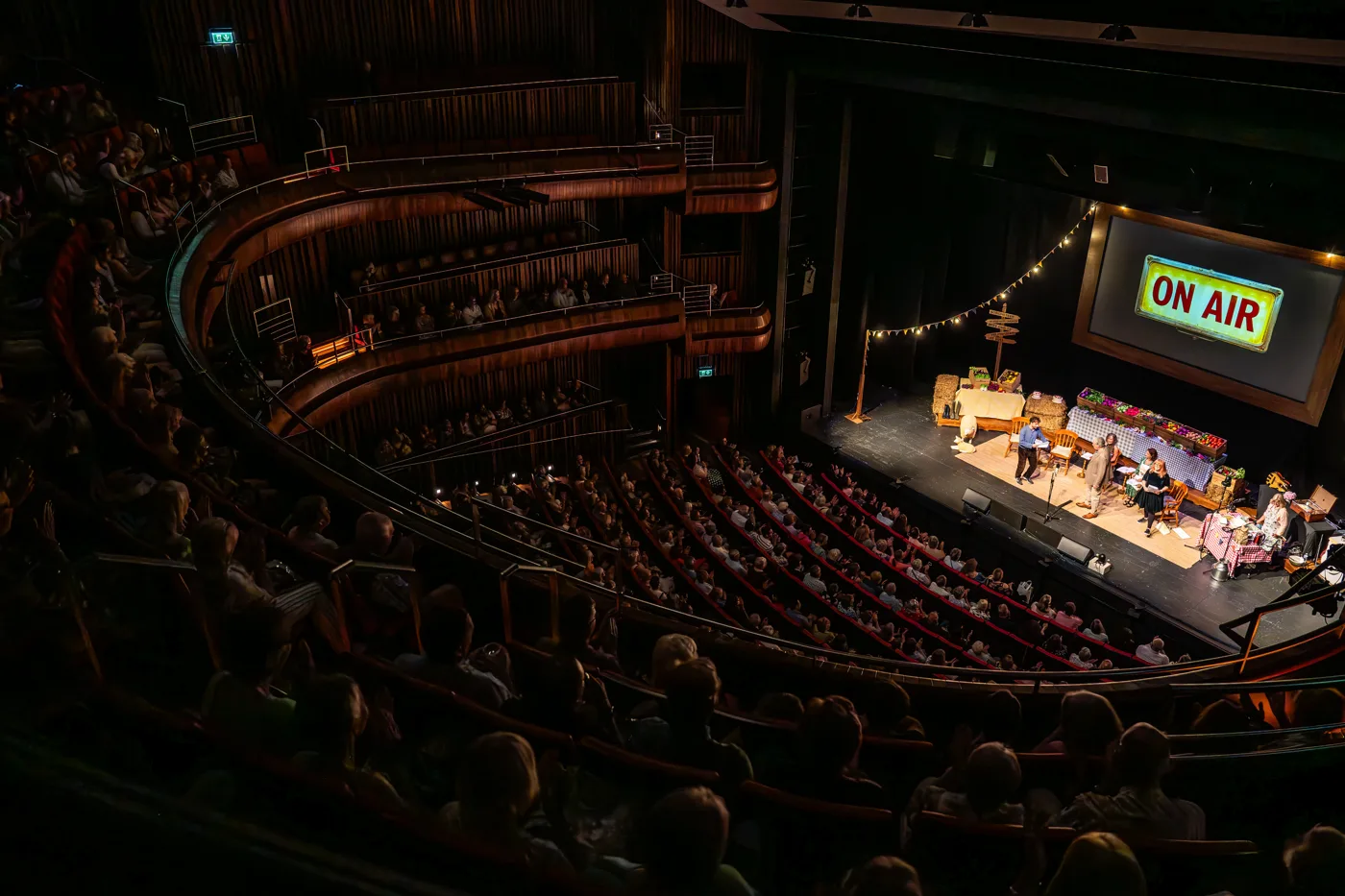 A large theatre auditorium seen from high in the circle, with an On Air sign lit above the stage