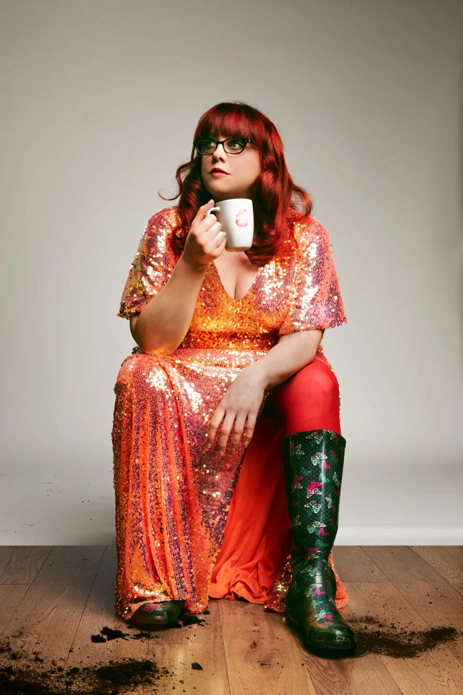 Angela Barnes in a sequinned orange dress and green wellies, sitting and holding a mug