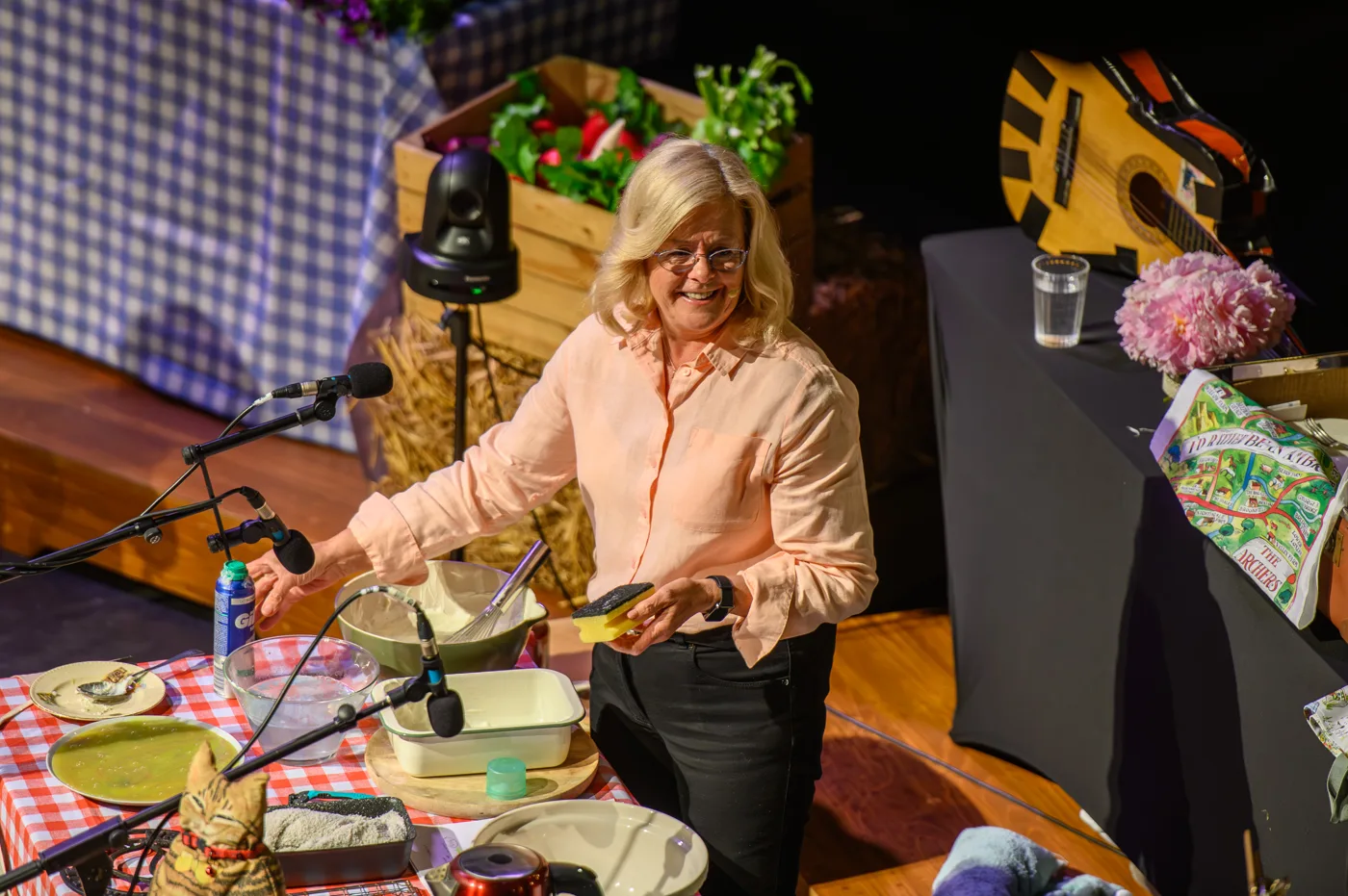 A woman at a table on stage covered with bowls, dishes and props, surrounded by microphones