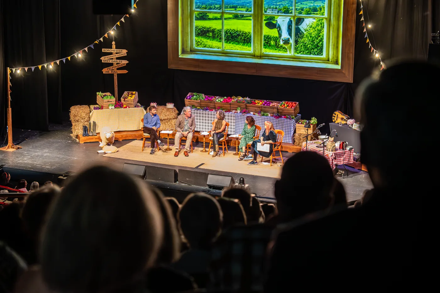 A theatre stage dressed as a village show, with bunting, a signpost, trestle tables of produce and a large window showing fields and a cow, seen past the heads of the audience
