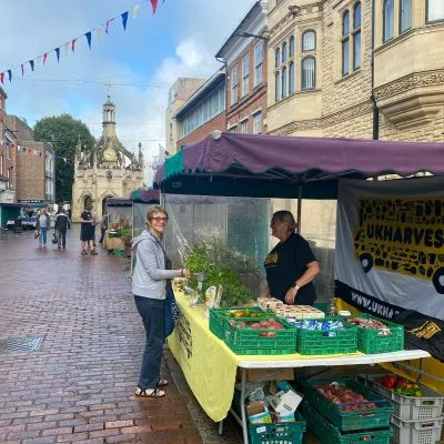 A UKHarvest stall with crates of vegetables and a yellow UKHarvest banner on a brick pavement in Chichester city centre, with the Market Cross in the background.